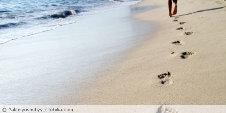 Person läuft Barfuß am Strand den Sand entlang, dabei entstehen Fußabdrücke.