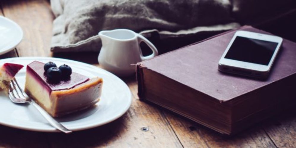 Auf dem Tisch steht ein Teller mit einem Kuchen, eine Milchkanne, auf dem Buch liegt ein Smartphone.