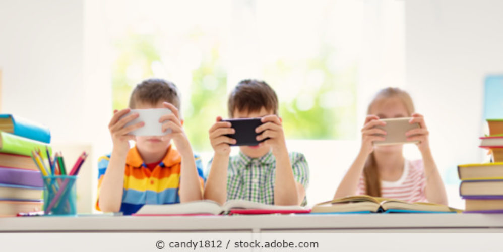 drei Kinder sitzen am Tisch und halten ihr Handy vor die Augen