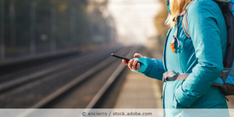 Frau steht an einem Bandgleis mit dem Handy in der Hand und wartet auf den Zug.