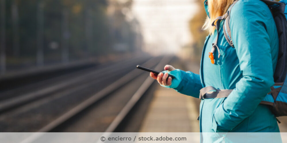 Frau steht an einem Bandgleis mit dem Handy in der Hand und wartet auf den Zug.