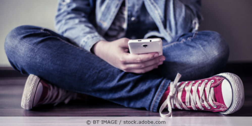 Teenager sitzt auf dem Boden und schaut auf sein Smartphone.