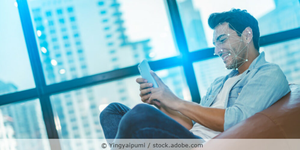 Junger Mann sitzt in einem Sessel und hält sein Smartphone in der Hand und ist über dem Gesicht der Person mit Punkten und Linien eine Gesichtserkennung als Fotomontage zu sehen.