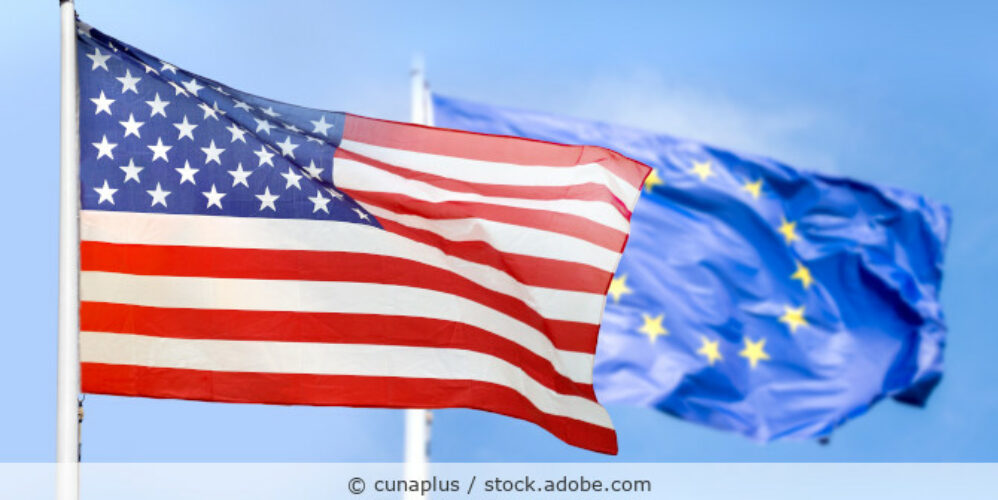 Zwei Flaggen, die der USA und der EU, bewegen sich im Wind vor blauem Himmel.