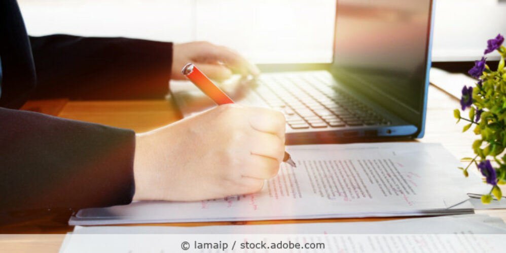 Frau sitzt am Schreibtisch vor einem Laptop und korrigiert einen ausgedruckten Text mit einem roten Stift.