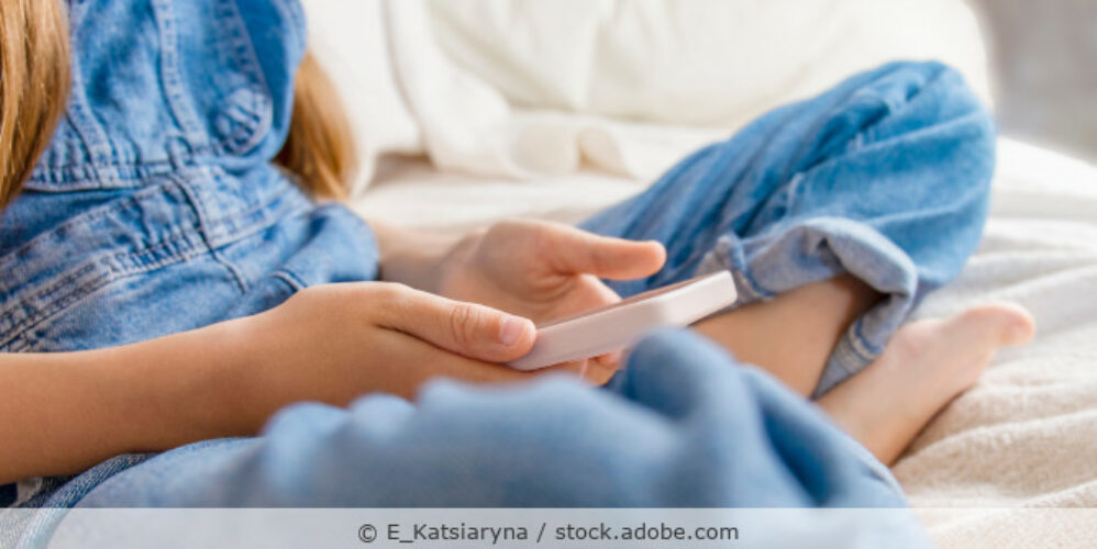 Kind mit Smartphone in der Hand sitzt auf dem Bett.