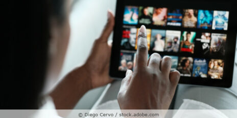 Eine Frau hält ein Tablet in der Hand, auf dem schemenhaft verschiedene Filme eines Streamingdienst-Anbieters zu sehen sind.