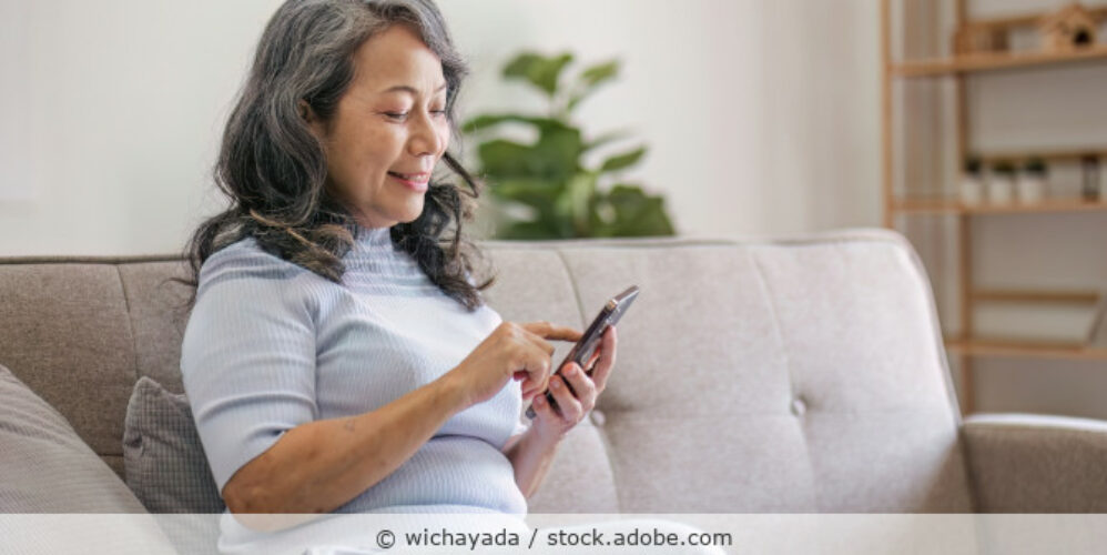 Eine etwa sechzigjährige Frau sitzt auf einem Sofa und hält ihr Smartphone in der Hand.
