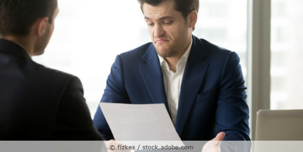 Zwei Geschäftspartner sitzen im Büro an einem Tisch, der eine hält einen Vertrag in der Hand und macht ein unzufriedenes Gesicht.
